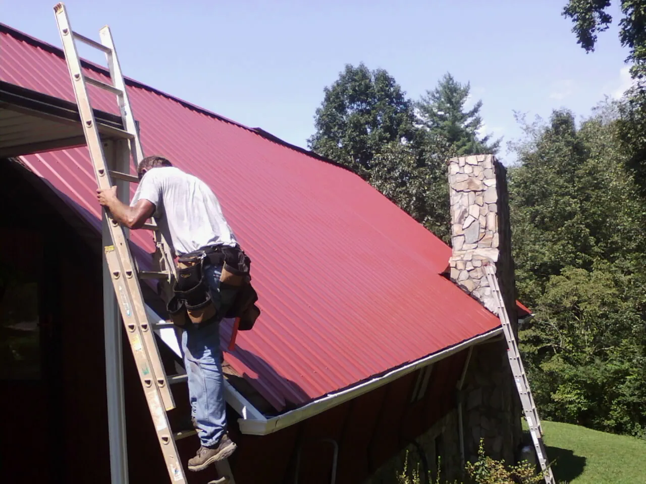 Roofer on a ladder beside a red metal roof and stone chimney — Soffit & Fascia Repair by Roofing Brothers in Ellendale